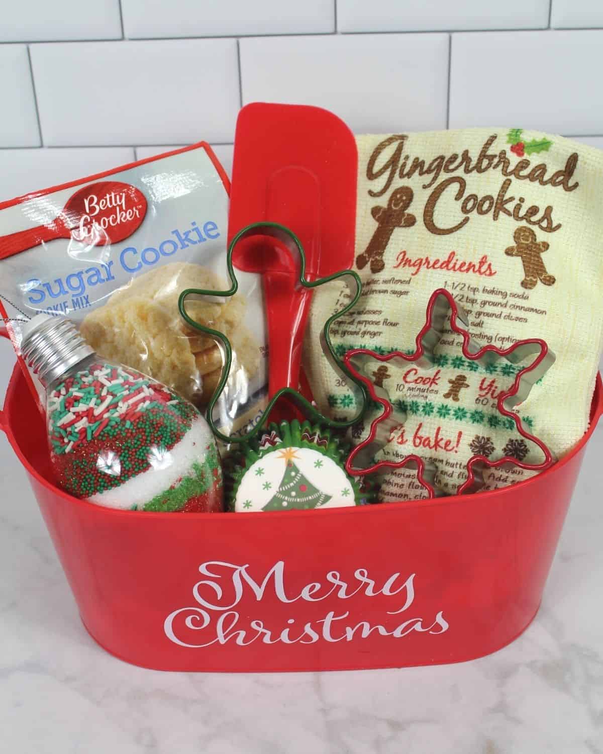 Dollar Tree holiday gift basket filled with baking essentials like Christmas cookie cutters, kitchen towel, clear ornament with sprinkles, Betty Crock sugar cookie mix, cupcake wrappers with Christmas trees on them and a red spatula all in red plastic gift basket that says "Merry Christmas" on a light grey marble background and subway tile backsplash