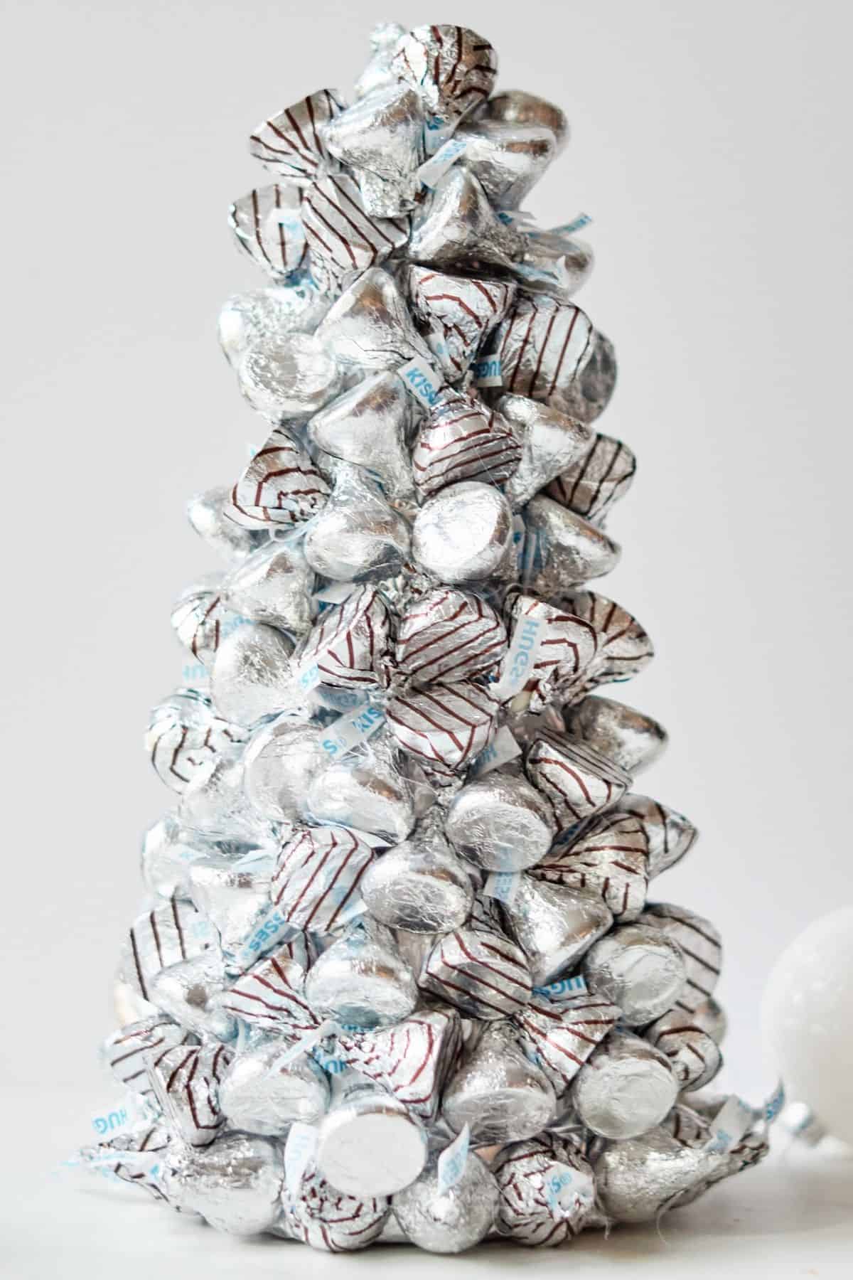 Hershey Christmas tree made of Hershey kisses and Hershey's Hugs candy on a grey background with a white holiday ornament in the background